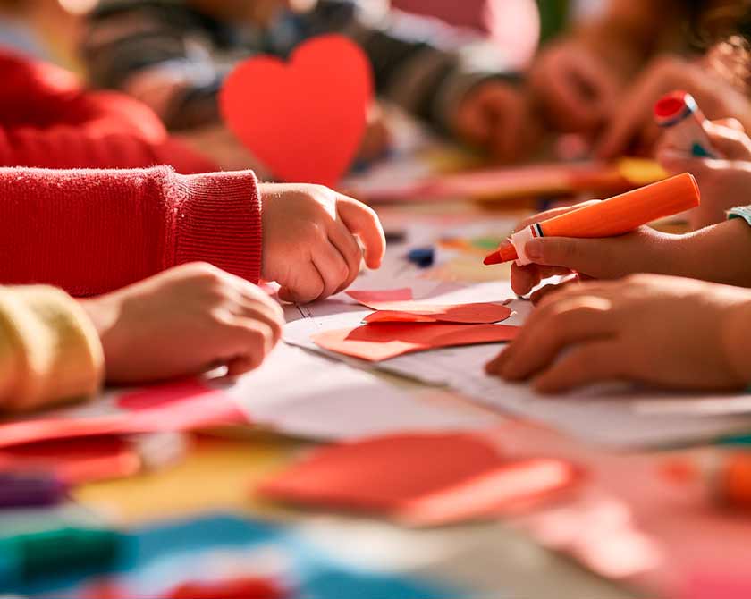 Manos de niños trabajan con marcadores y corazones de papel sobre una mesa de aula durante una actividad grupal
