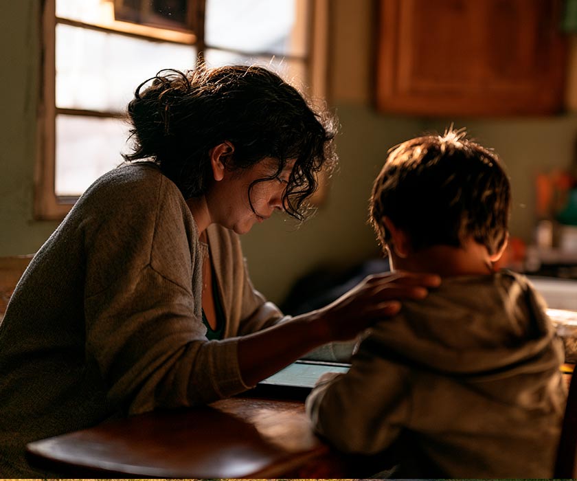 Imagen ilustrativa: una madre acompaña a su hijo mientras usa una tableta en la mesa de la cocina de su casa.