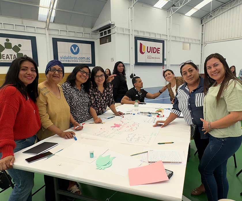 Un grupo de docentes sonríe alrededor de una mesa con papelógrafos y marcadores durante un taller de capacitación.